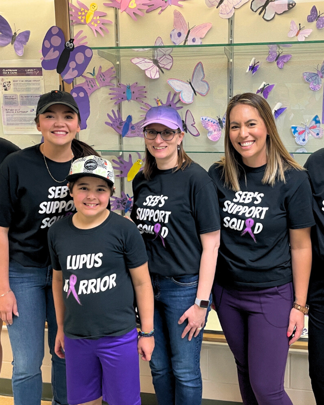 Sebastian, a young lupus patient, stands smiling with his teachers in front of a wall of purple butterflies made by students to raise awareness for lupus.