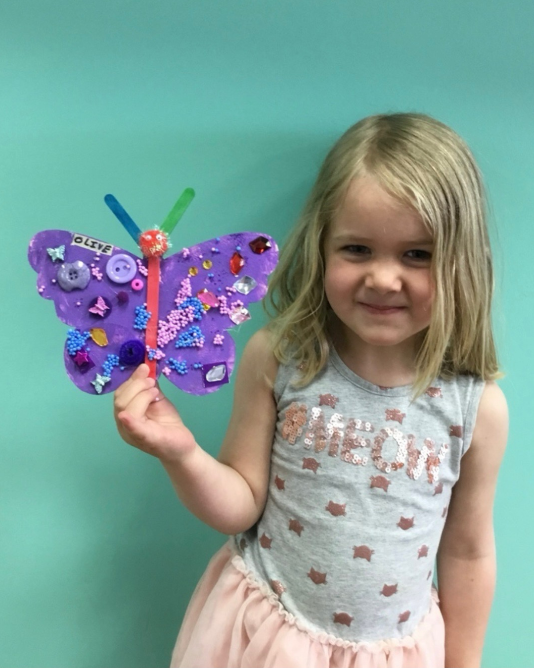A young girl smiles while holding a decorated purple butterfly craft at Rising Star Preschool and Daycare.