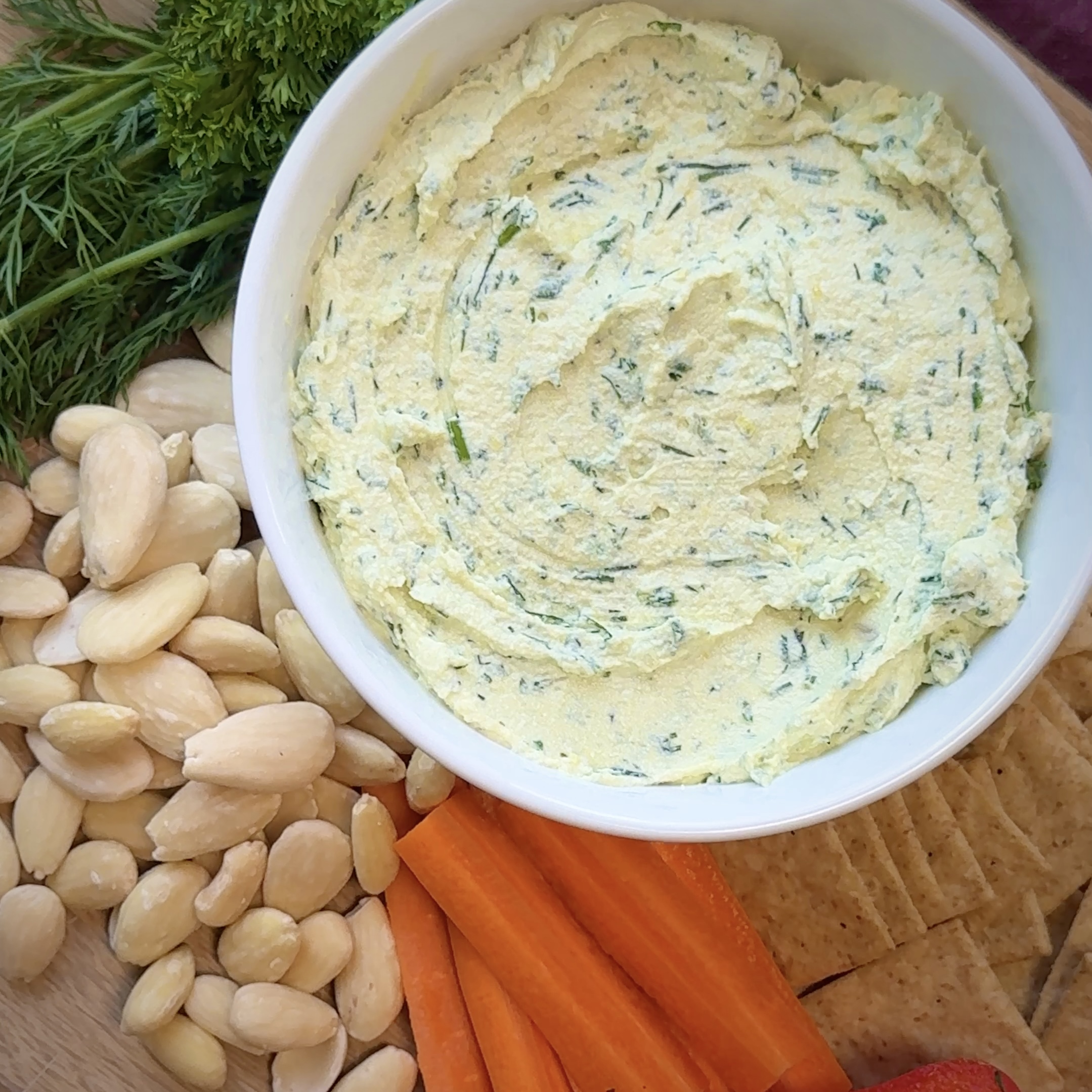 Un bol de trempette crémeuse au tofu aux herbes entourée d’amandes blanchies, d’aneth frais, de persil, de bâtonnets de carottes, de craquelins et de tranches de pomme.