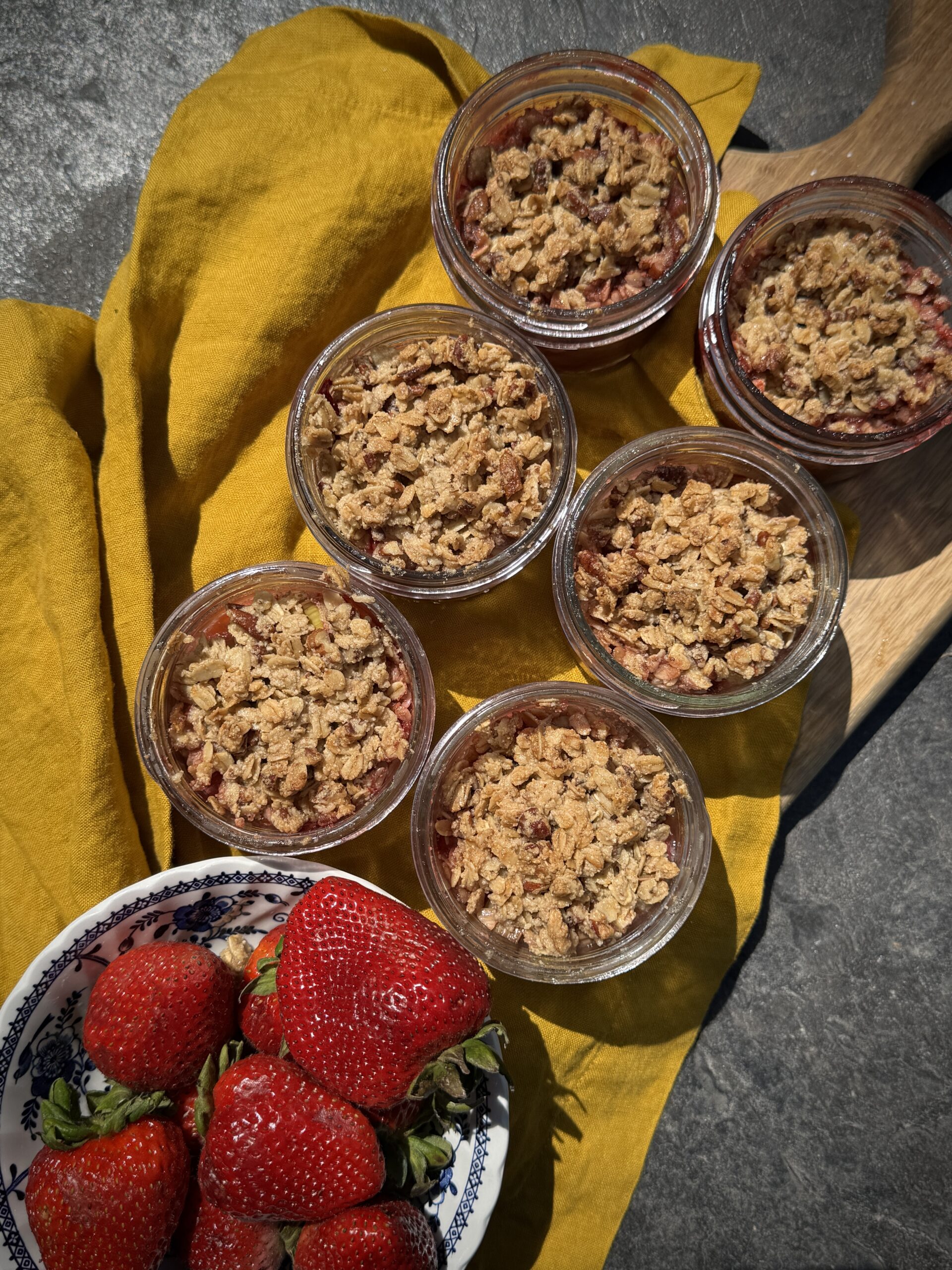 Overhead view of individual gluten free strawberry rhubarb crumbles in mason jars, placed on a yellow cloth with fresh strawberries in a bowl.