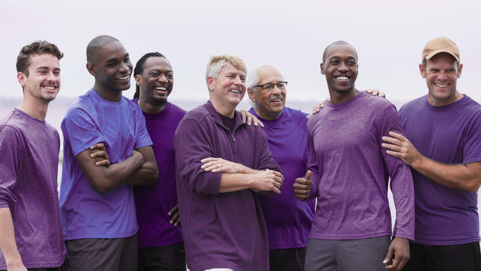 Men and lupus of various ages standing together outdoors, all wearing different shades of purple and smiling.