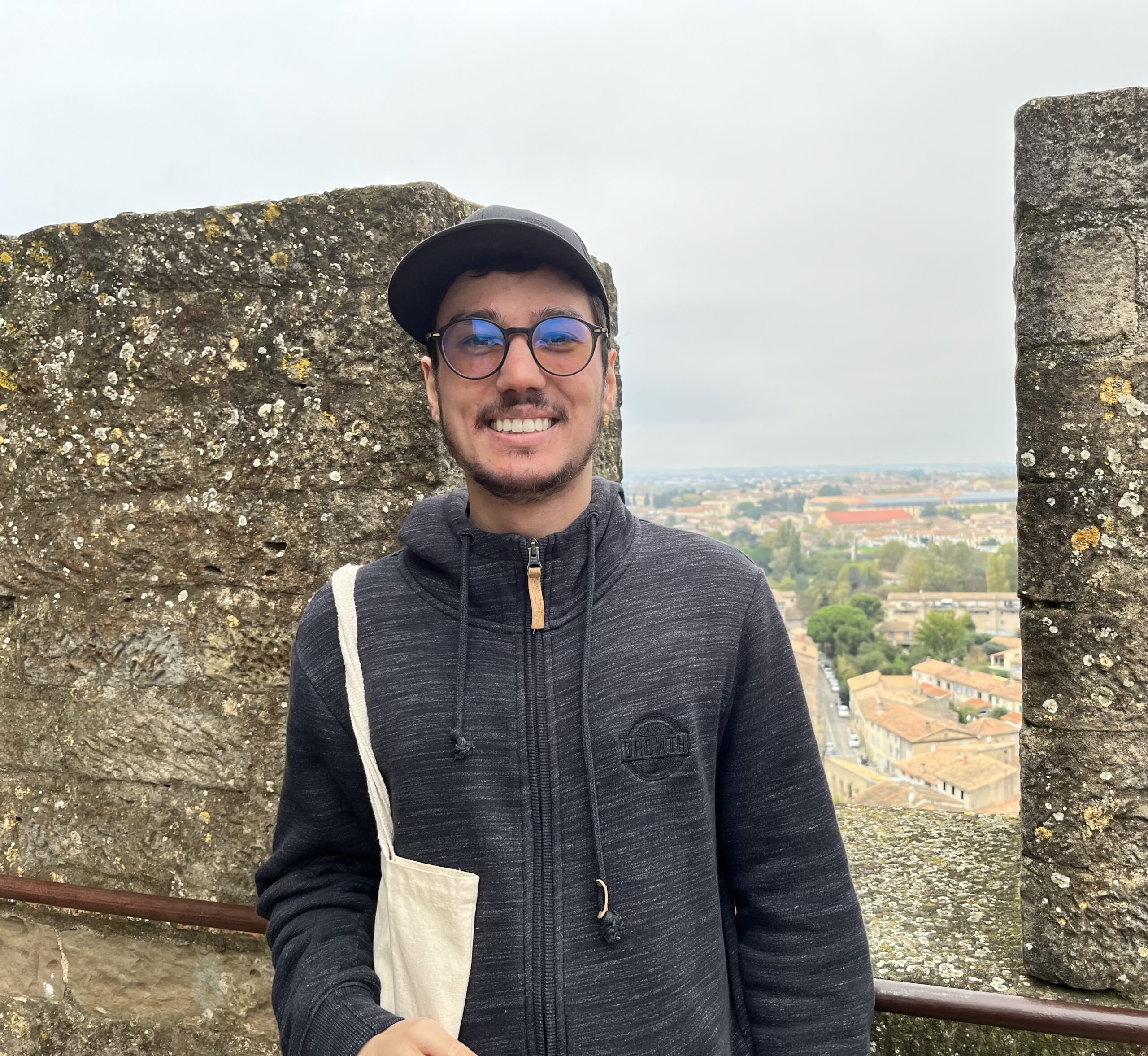 Jonathan standing on a stone balcony, smiling, with a panoramic view of rooftops and a cloudy sky in the background