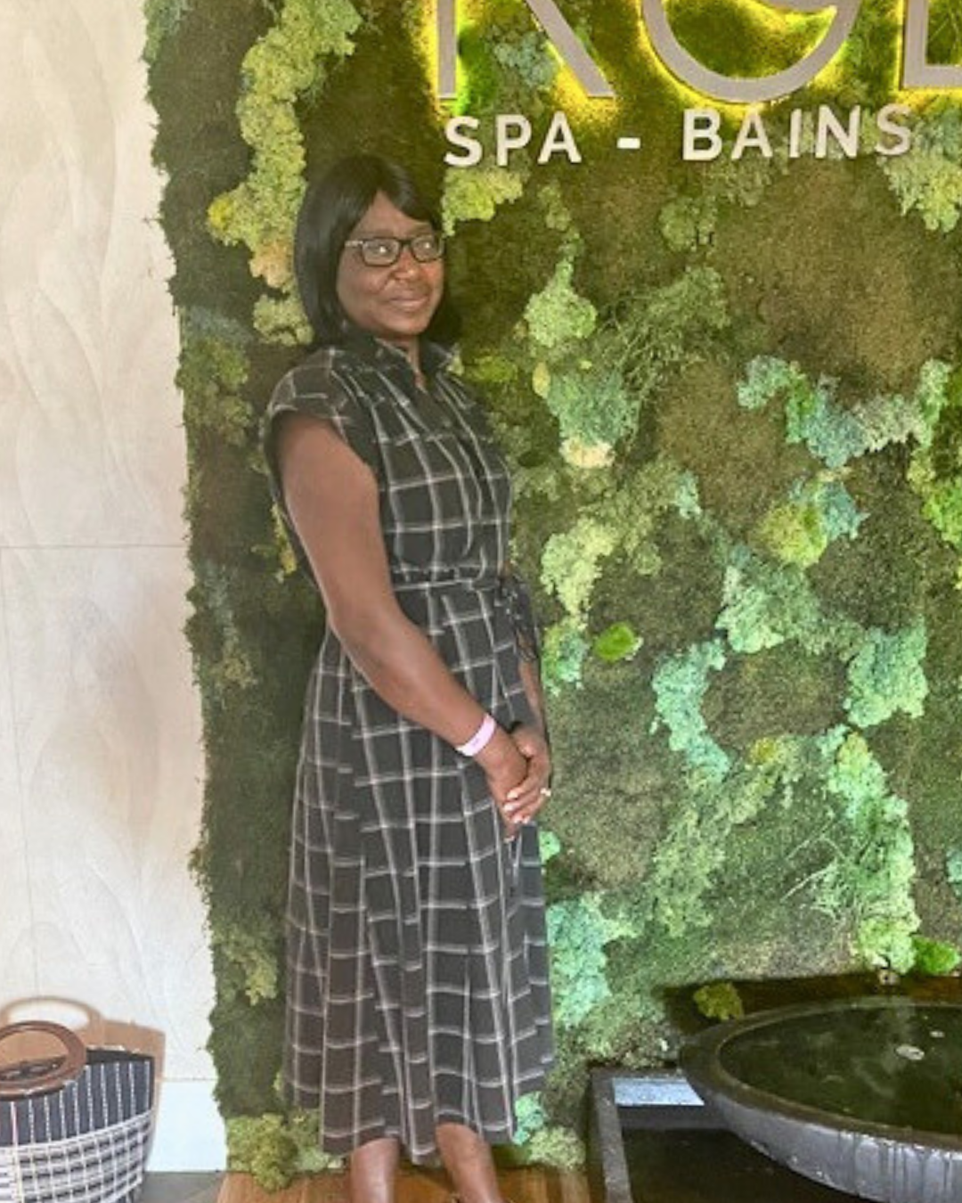 Anita Toni standing in front of a moss-covered spa wall, smiling gently and wearing a black patterned dress and colorful sandals.