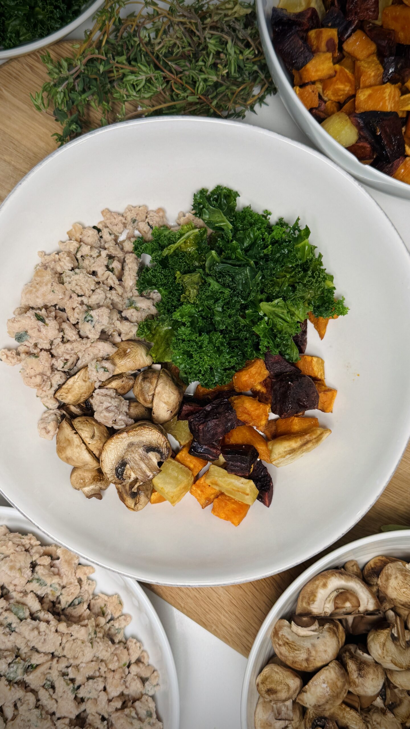 Bowl of Chicken Sausage Hash featuring ground chicken, sautéed mushrooms, roasted yams and root vegetables, and steamed kale, surrounded by fresh ingredients like herbs and mushrooms