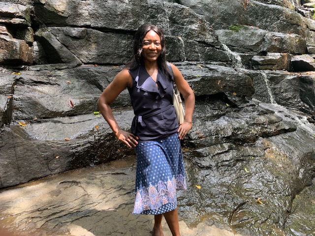 Anita Toni standing in front of a rocky stream on a sunny day, smiling confidently with her hands on her hips