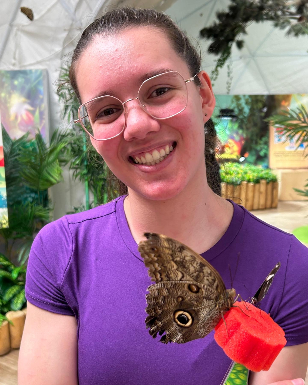 Yaneisy Nynoska Tremblay souriante dans une serre de papillons, tenant un grand papillon brun avec des taches oculaires sur une mangeoire rouge, Vivant avec le lupus.