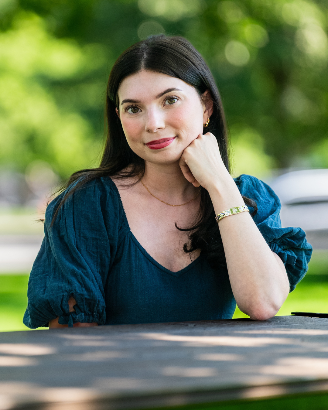 Personne aux cheveux noirs assise à une table de pique-dans un parc verdoyant, reposant son menton sur une main et souriant à la caméra.
