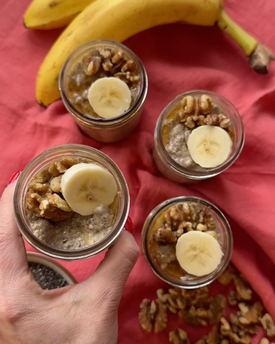 Banana coconut chia pudding in small jars topped with sliced banana and walnuts, styled on a red cloth with fresh bananas.
