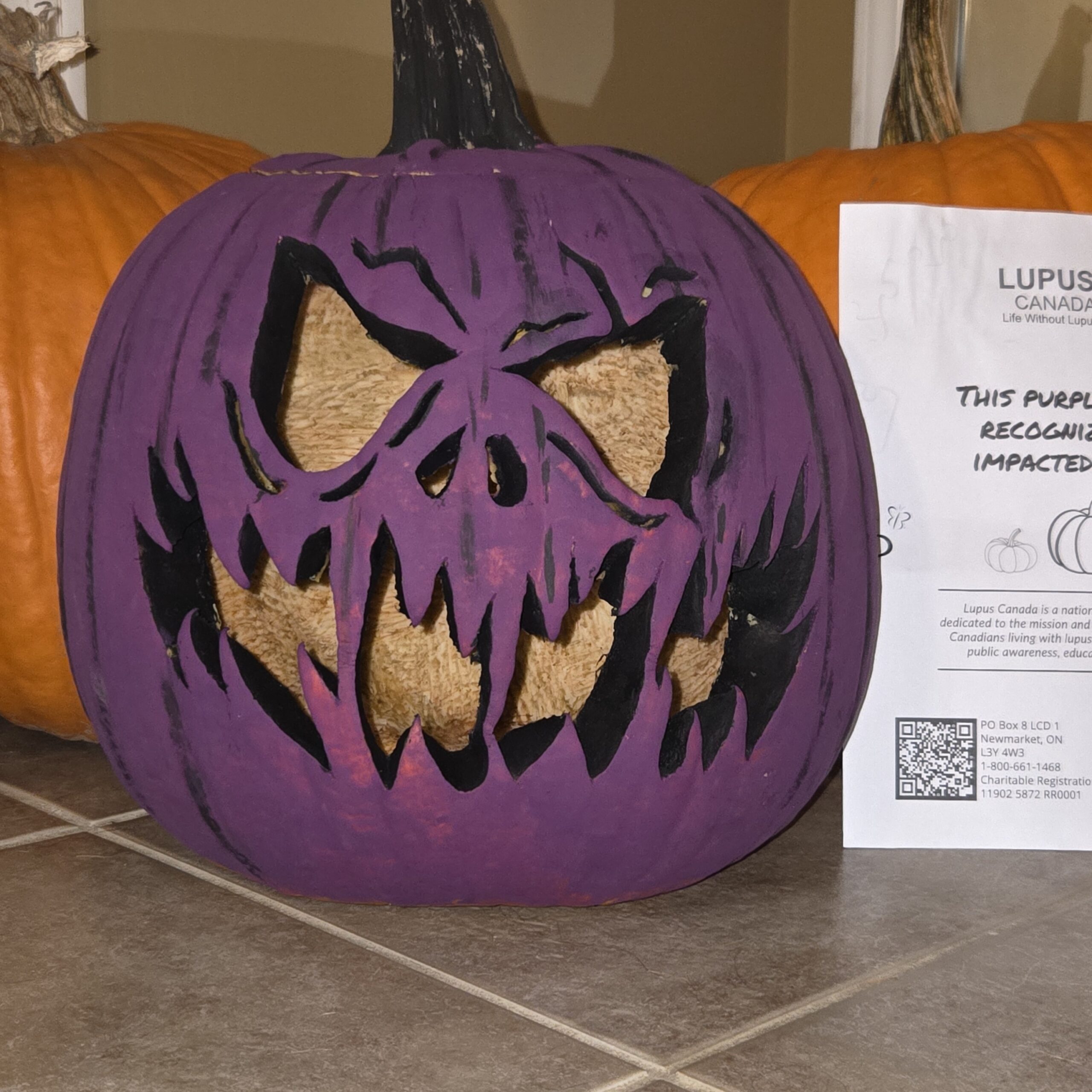 Une citrouille violette sculptée avec un visage audacieux et dentelé est placée à côté d'une affiche de Lupus Canada sur laquelle on peut lire : « Cette citrouille violette rend hommage aux personnes touchées par le lupus. »