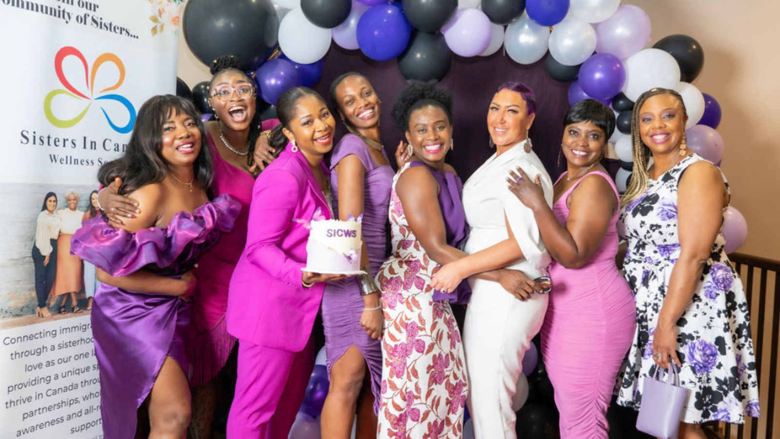 Groupe de femmes vêtues de violet lors d’un événement communautaire de bien-être, debout ensemble devant un présentoir de ballons et une bannière Sisters in Care