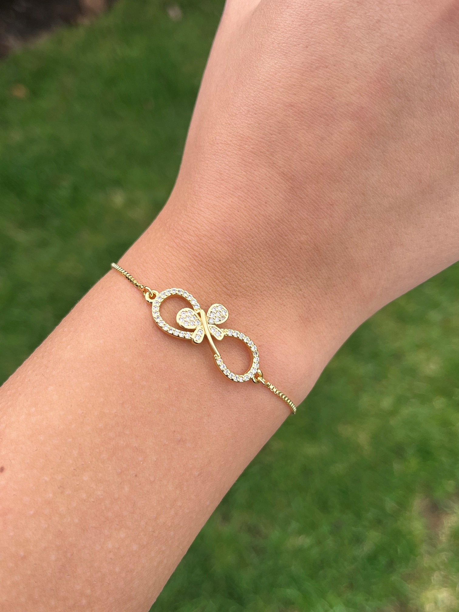 Close up of a gold filled infinity bracelet with a butterfly detail and sparkling CZ stones worn on a wrist outdoors. The Infinity of Hope bracelet was created in partnership with Two Charmed to support lupus awareness and research through Lupus Canada.