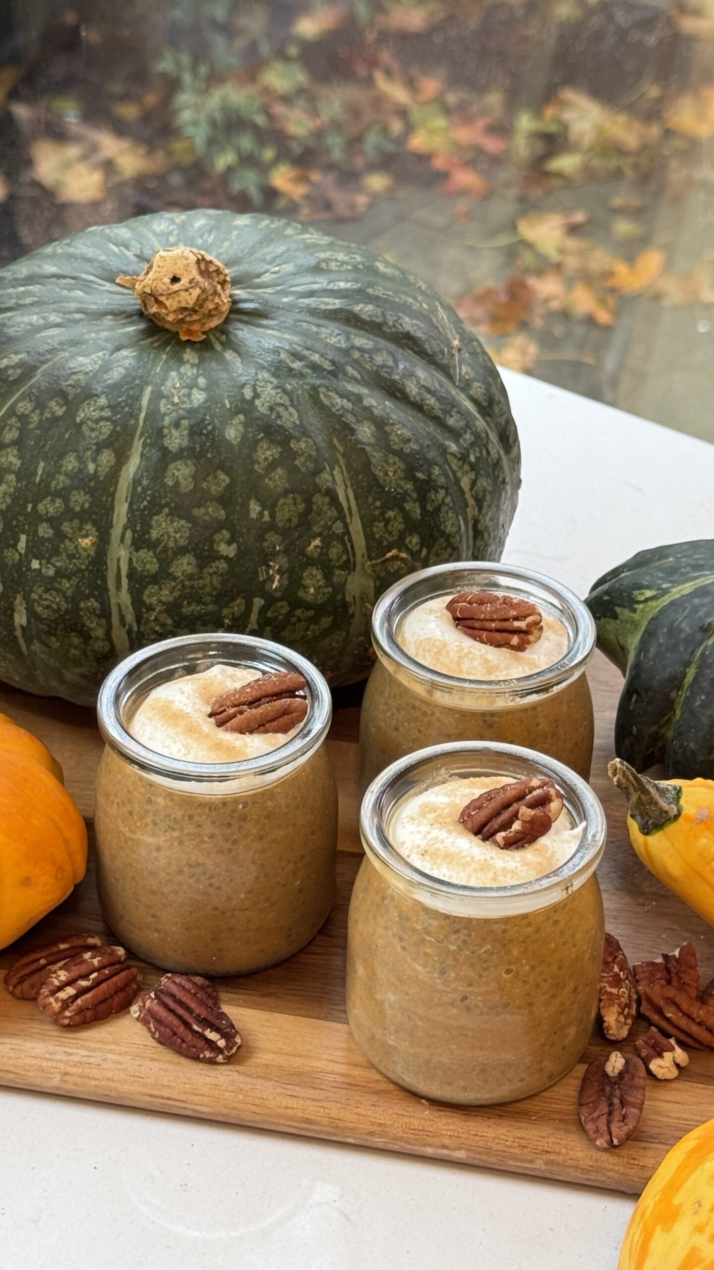 Trois bocaux en verre de pudding au chia à la citrouille garnis de crème de coco et de pacanes, entourés de citrouilles et de décorations d’automne.